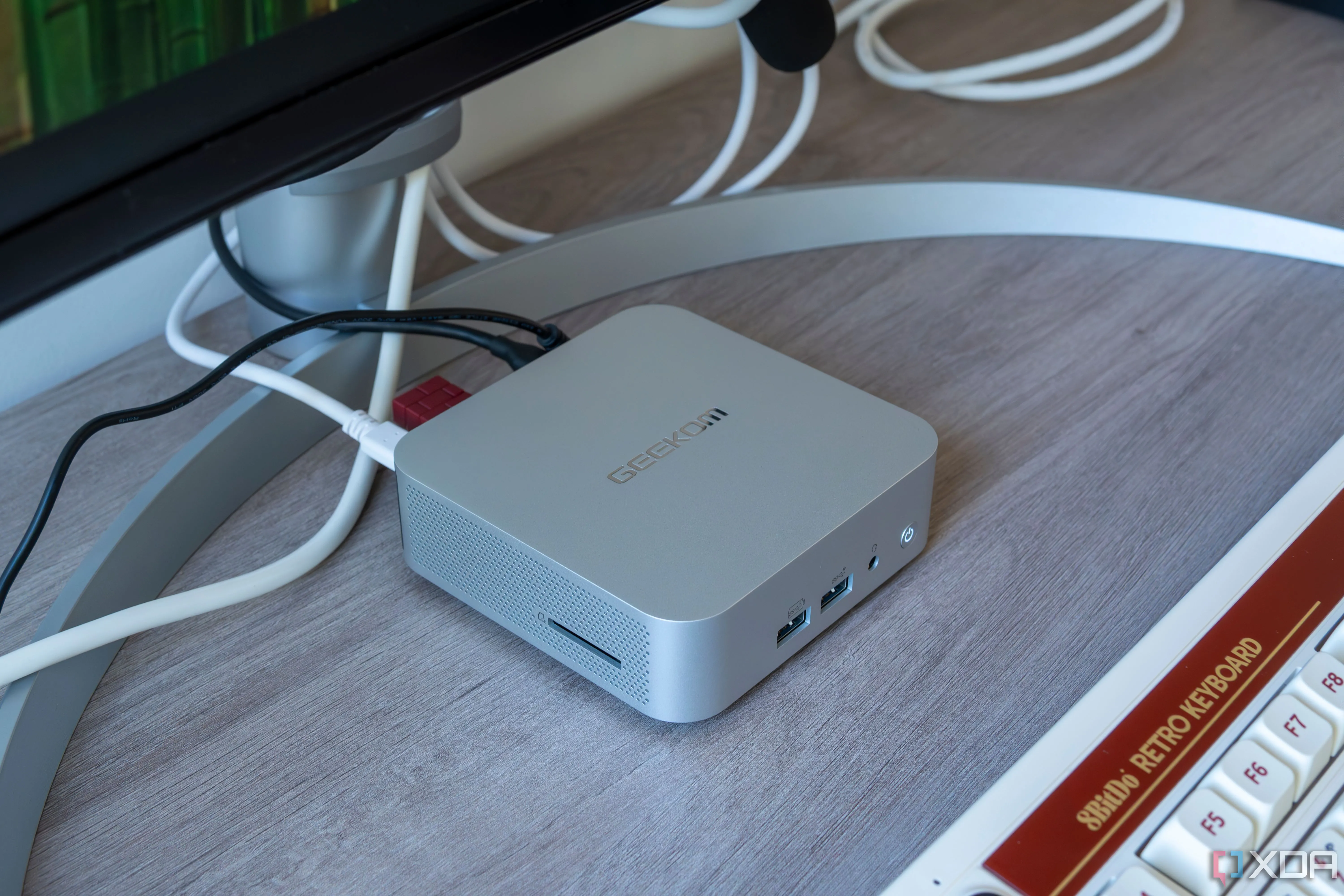 Mini PC Geekom A8 đặt giữa màn hình và bàn phím, làm nổi bật các cổng kết nối và thiết kế nhỏ gọn